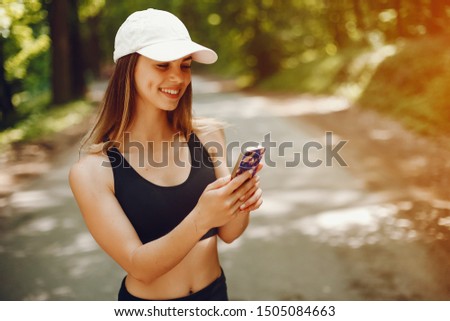 Cute girl training in a summer forest. Lady have fun in a park. Sports woman in a black sportsuit. Famale in a white cap use the phone