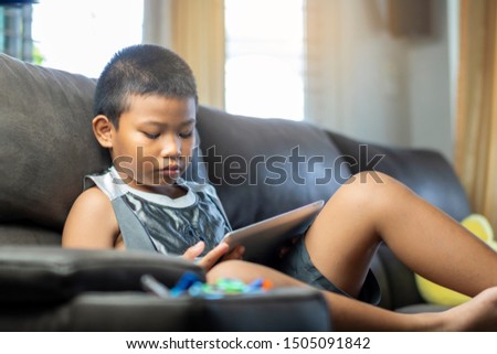 Asian boy playing game or watching cartoons on digital taplet or smartphone on sofa living room.