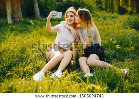 two beautiful young beautiful girls with shiny blond hair and a skirt and sitiing with phone in a sunny summer park