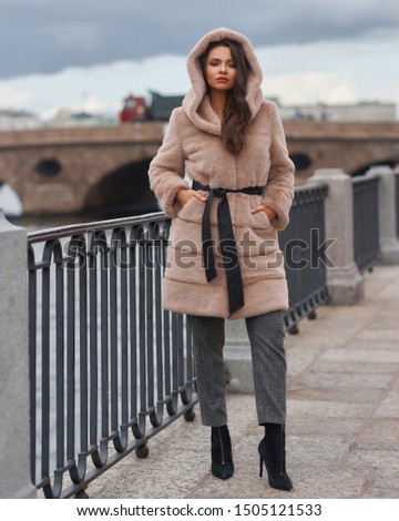 Young gorgeous woman with long wavy brunette hair in beigr fur coat standing and posing at city embenkment. Rich expensive woman. Full length portrait