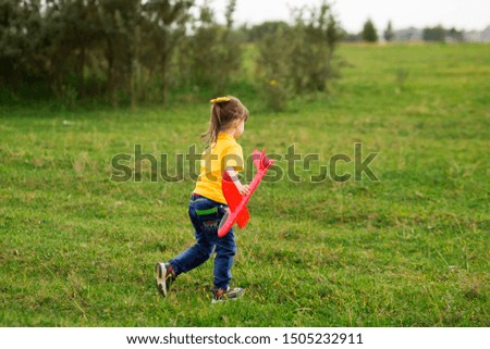 A preschooler girl in a yellow T-shirt and jeans runs forward to the right on the grass with a red toy airplane. summer or autumn, evening. horizontal photograph of a child.