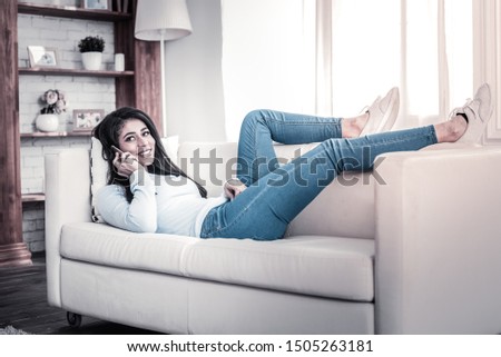 Lying on sofa. Beautiful long-haired girl having conversation on her phone and openly smiling during it