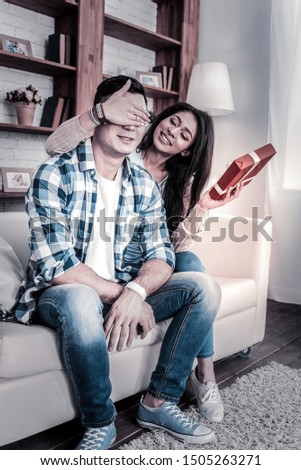 Man closing eyes. Appealing lady closing eyes of her husband and carrying his present in red cover in other hand