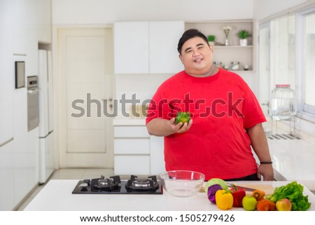 Picture of happy fat man holding a green paprika while preparing to make a healthy salad in the kitchen