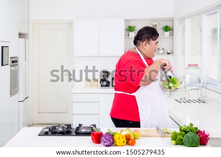 Picture of a young fat man wearing an apron while preparing a bowl of organic salad in the kitchen