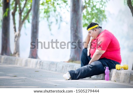 Picture of young obese man looks unhappy after exercise while sitting in the park