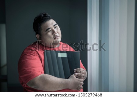Picture of an obese man looks unhappy while holding a weight scale and standing near the window