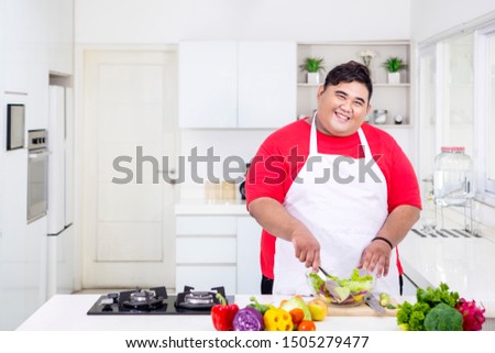 Picture of smiling fat man mixing a tasty salad while standing in the kitchen