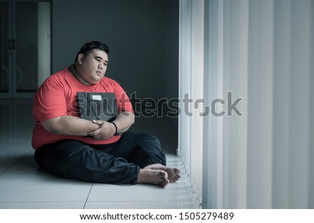 Picture of an unhappy fat man holding a weight scale while sitting near the window