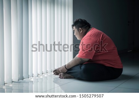 Picture of young fat man looks pensive while sitting near the window 