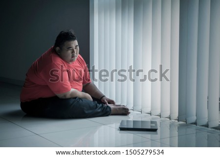Picture of overweight man looking at the weight scale while sitting near the window 