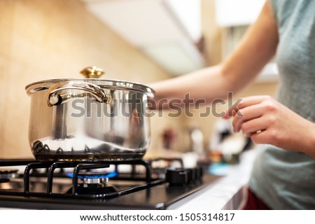 Beautiful young woman housewife prepairing dinner, hold in hands big steel saucepan, standing it on gas-stove. Royalty-Free Stock Photo #1505314817
