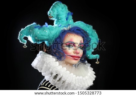 girl in makeup and costume jester . clown girl with bright makeup in blue wig On black background. She looks naughtily at the camera 