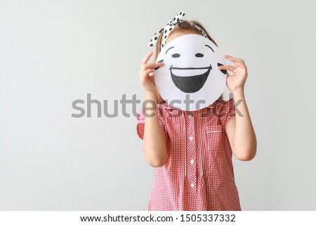 Little girl hiding face behind drawn emoticon on light background