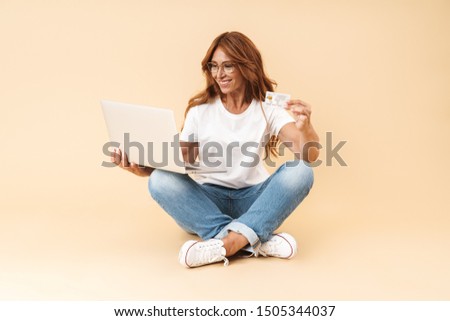 Happy middle aged woman wearing casual outfit sitting on a floor with legs crossed isolated over beige background, showing plastic credit card while using laptop computer