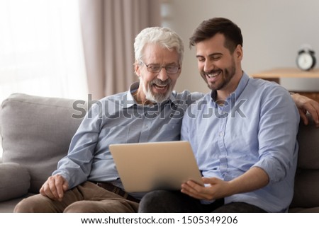 Happy two age generation family senior father and adult son having fun enjoy using laptop at home sit on couch, laughing old dad with young man bonding watching funny video looking at computer screen