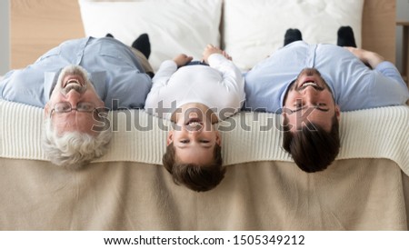 Happy multi 3 three generation family lying upside down in row on bed looking at camera, smiling child boy son grandson, young father and old grandfather bonding posing for funny portrait in bedroom