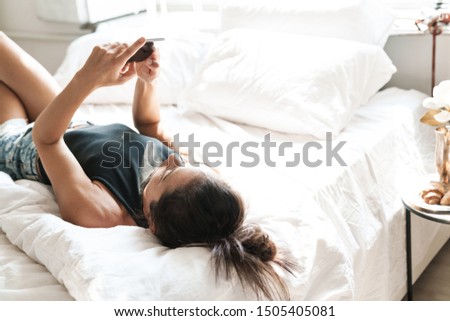 Image of a young beautiful woman indoors at home on bed using mobile phone.