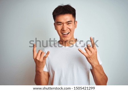 Young asian chinese man wearing t-shirt standing over isolated white background shouting with crazy expression doing rock symbol with hands up. Music star. Heavy music concept.