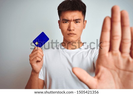 Young asian chinese man holding credit card standing over isolated white background with open hand doing stop sign with serious and confident expression, defense gesture