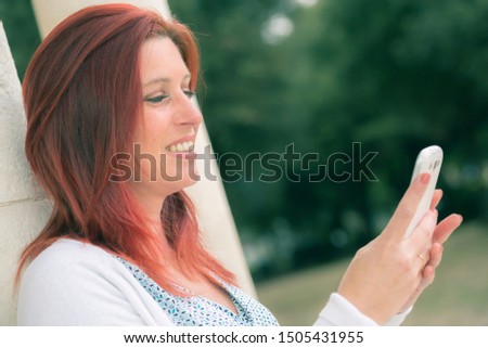 Pretty smiling redhead woman sending messages or make a selfie on her cell phone in a park