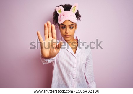 Young african american woman wearing pajama and mask over isolated pink background doing stop sing with palm of the hand. Warning expression with negative and serious gesture on the face.