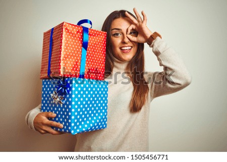 Young beautiful woman holding birthday gifts standing over isolated white background with happy face smiling doing ok sign with hand on eye looking through fingers