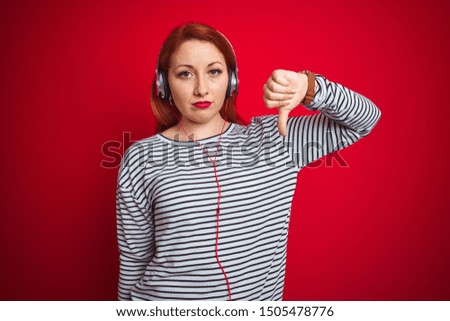 Young redhead woman listening to music using headphones over red isolated background with angry face, negative sign showing dislike with thumbs down, rejection concept