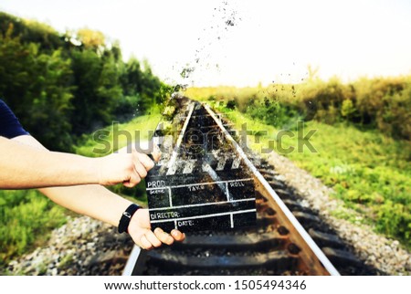 Guy is holding black clapperboard falling into pieces. Man is directing and filming some amateur cinema movie. Mysterious image. Rail trails on the background. Travel concept.