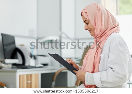 Side view portrait of Middle-Eastern woman wearing hijab working as nurse in medical clinic and writing on clipboard, copy space