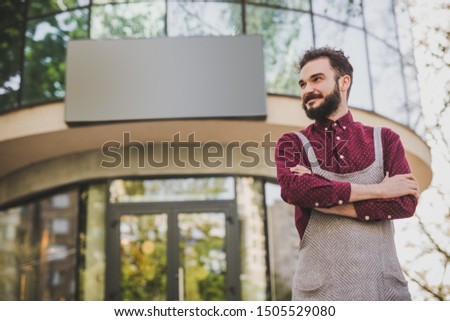 Low angle of positive bearded man in apron smiling and looking away while standing with crossed arms outside modern restaurant building with empty banner