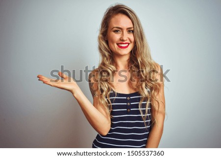 Young beautiful woman wearing stripes t-shirt standing over white isolated background smiling cheerful presenting and pointing with palm of hand looking at the camera.