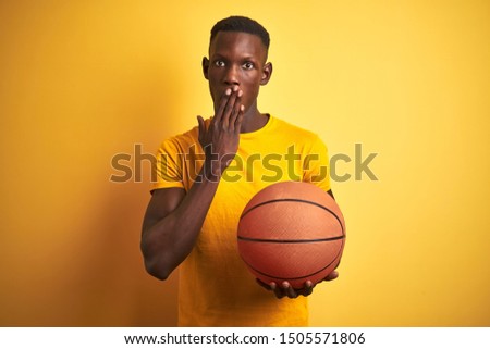 African american athlete man holding basketball ball standing over isolated yellow background cover mouth with hand shocked with shame for mistake, expression of fear, scared in silence
