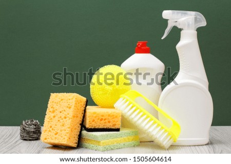 Plastic bottles of dishwashing liquid, glass and tile cleaner, yellow brush and sponges on laminate flooring and green background. Washing and cleaning concept.