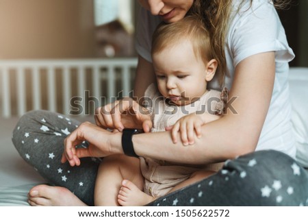 Nine-month-old baby girl with her mother is sitting on bed and looking at screen of smart watch on her mother s hand. In background crib in soft focus. Mom and her daughter watch time on wristwatch. Royalty-Free Stock Photo #1505622572