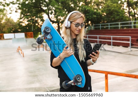Photo of caucasian pleased woman wearing headphones and sunglasses typing on cellphone while holding board in skatepark
