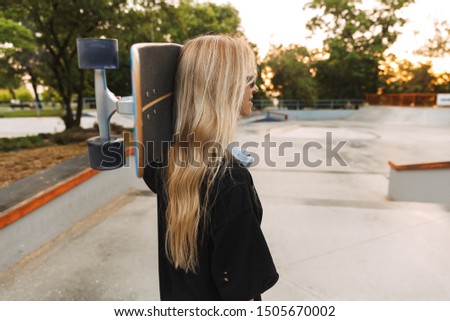 Attractive funky teenage girl carrying longboard while walking at the skatepark