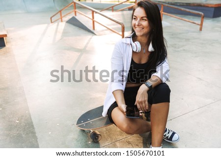 Pretty young teenage girl sitting at the skatepark with a longboard, holding mobile phone