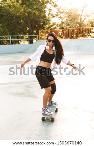 Photo of attractive young woman in streetwear wearing headphones riding skateboard in skate park