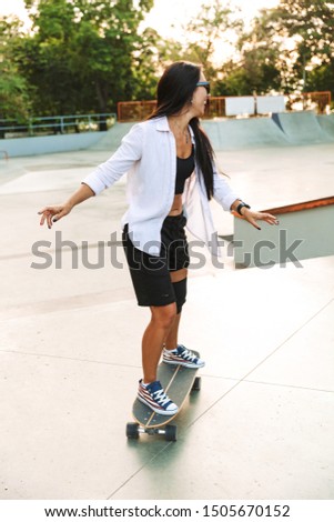 Photo of pretty young woman in streetwear smiling while riding skateboard in skate park