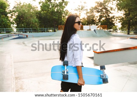 Photo of hipster young woman in streetwear walking and carrying skateboard in park