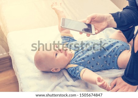 Side view of lying infant photographed by woman with smart phone. Child is stretching his arms and looks at the phone. All potential trademarks are removed. 