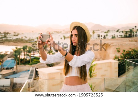 Young woman in straw hat and cute summer dress standing at the balcony with picturesque sea scenery, taking picture of sunset or sunrise on smartphone, back view