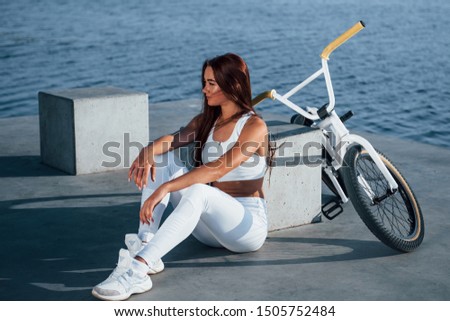 Near the bike. Fitness woman having a rest near the lake at daytime. Beautiful sunlight.