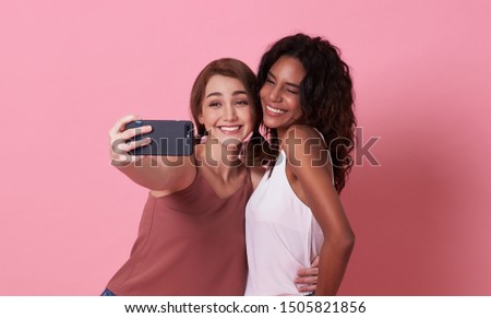 Two cheerful girls taking a selfie while showing gesture together over pink background.