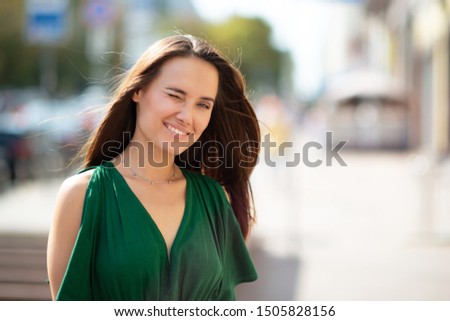 Young pretty likable cheerful woman posing summer city outdoor. Beautiful self-confident girl dressed in emerald-colored jumpsuit with long brown hair walking street enjoing her life, urban lifestyle