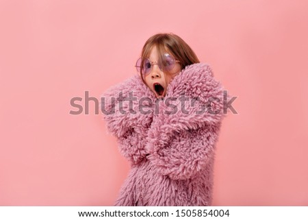 Close up portrait of lovely funny little girl wearing round violet glasses and stylish fur coat cover face with hands