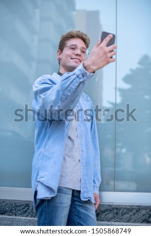 Concept of technological youth. Bottom view of a handsome smiling man with glasses and headphones taking a picture of himself with his smart cell phone. Portrait