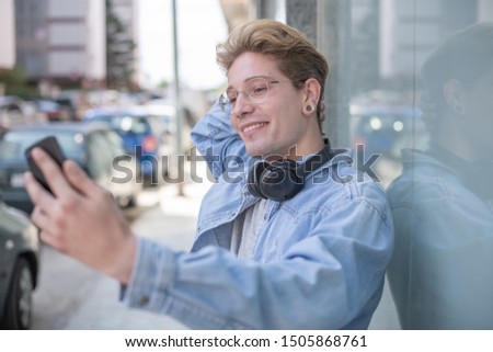 Portrait of a blond boy with glasses and headphones taking a picture of himself with his smartphone in the street. Concept of technological youth. Close-up