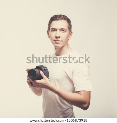 Serious Man with camera isolated on light background. Young man holding digital camera and look on you. Lifestyle, travel and technology concept. Happy boy in white t-shirt love work and dslr camera